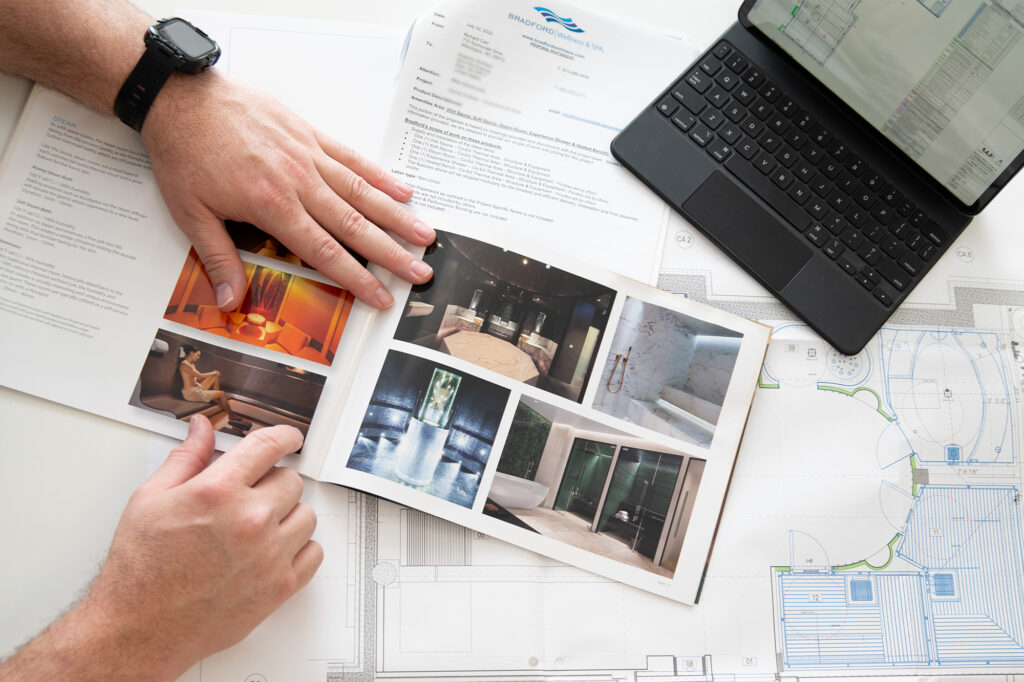 hands turning the pages of a bradford wellness & spa brochure. a laptop and spa design plans are also visible